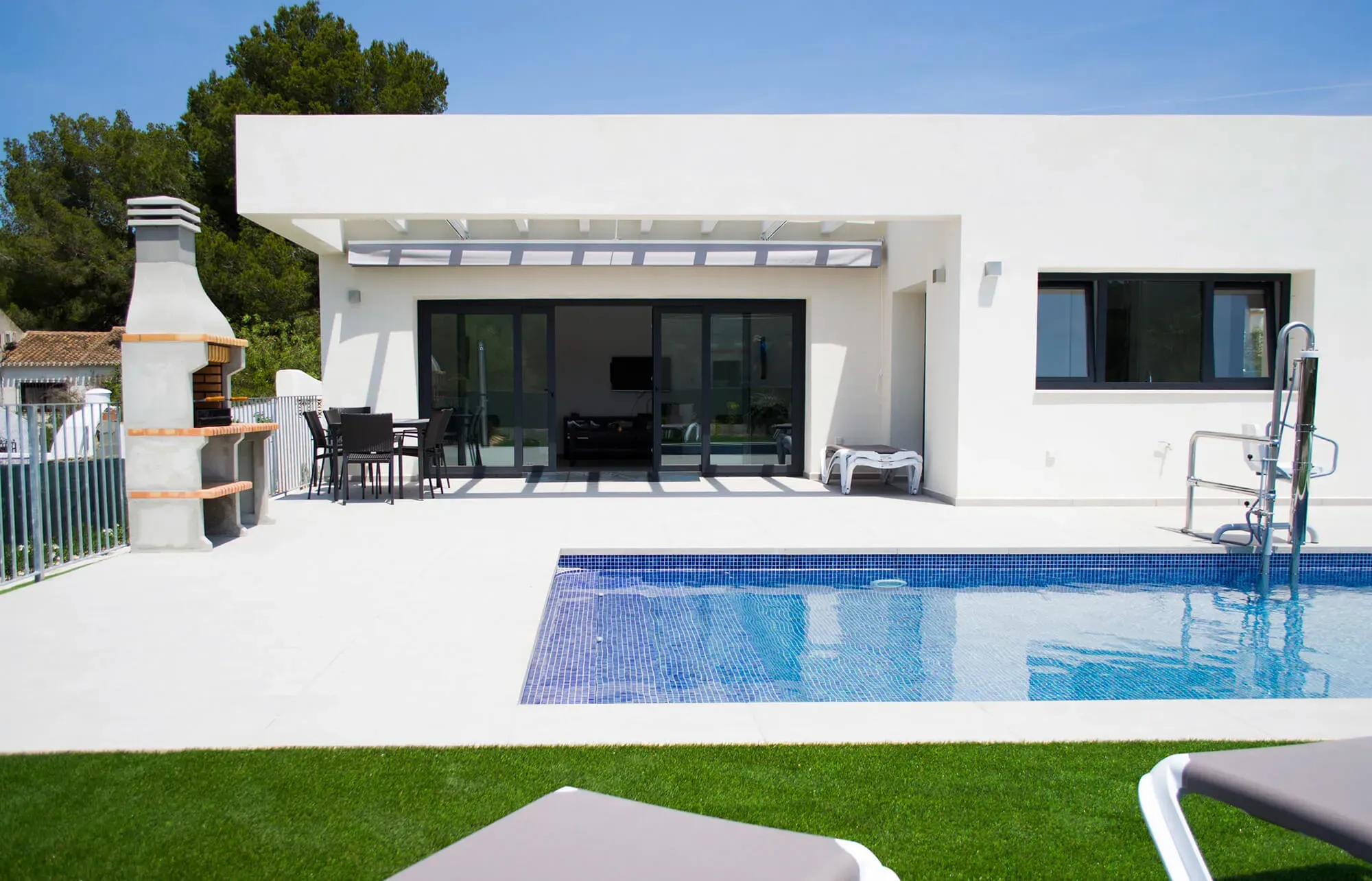 Villa Olivo from the garden: the white single-storey villa, its shaded terrace and the pool in the sun, with the pool hoist at the water's edge